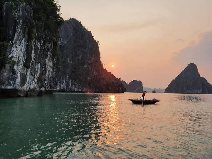A lone rower glides past towering limestone cliffs in Halong Bay at sunset.