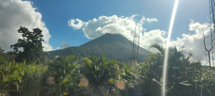 Conical Arenal Volcano rising above jungle vegetation under a bright sky with scattered clouds