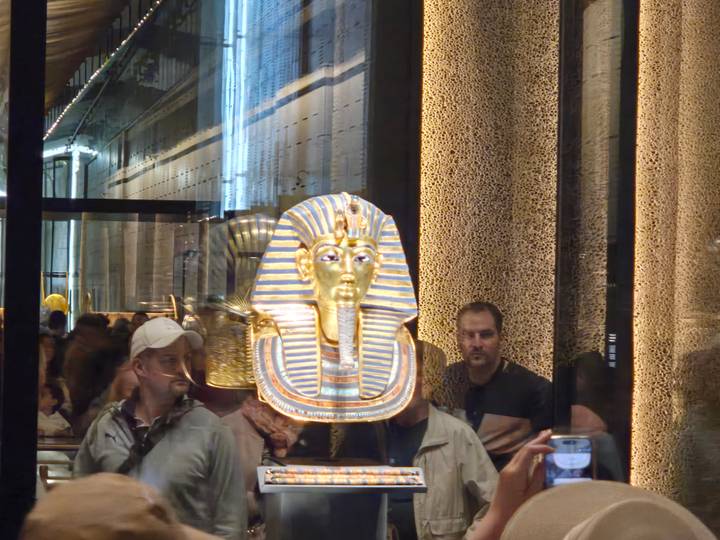 Golden mask of Tutankhamun behind glass inside the Egyptian Museum, with visitors looking on