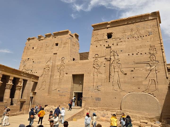 Sandstone walls of Philae Temple covered in hieroglyphs under a blue sky, visitors entering through central doorway