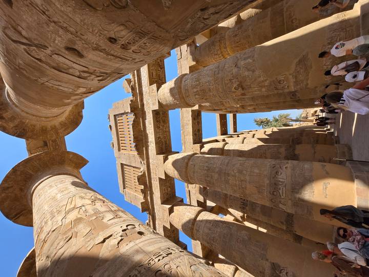 Upward view through the hypostyle hall of Karnak Temple showing massive carved sandstone columns against a blue sky
