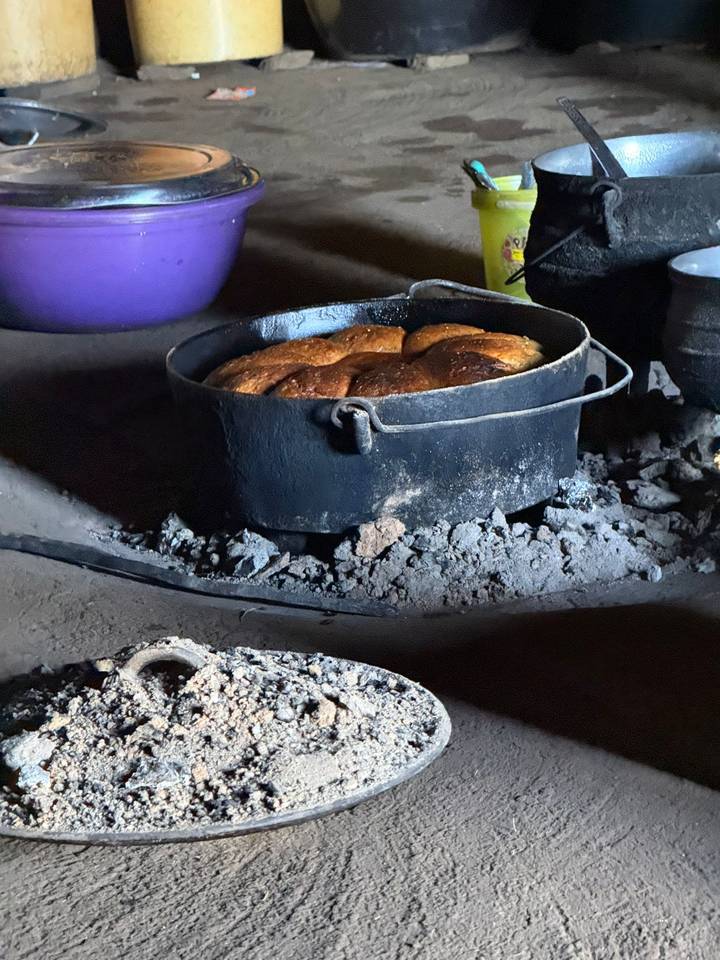Loaf of bread baking in a black cast-iron pot on hot embers inside a rustic hut.