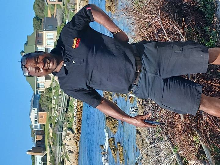 Smiling guide in front of seaside homes and rocky shoreline where penguins roam.