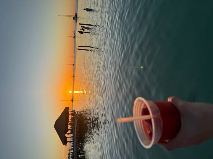 Tropical sunset over calm sea with sailboats while holding a fruit cocktail in foreground
