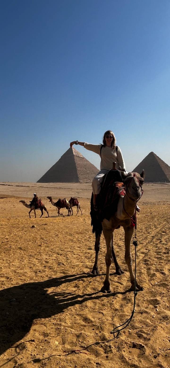 Woman on camel playfully posing as if pinching the Great Pyramid while other camels trek across the Giza Plateau.