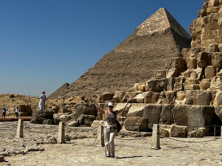 A visitor raises her arms in front of the Great Pyramid while another tourist stands on nearby rocks under a clear blue sky.