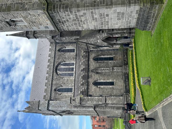 Gothic stone cathedral stands beside manicured lawns and blooming yellow flowers below a partly cloudy sky.