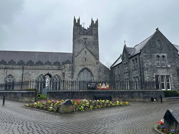Grey-stone church and surrounding railings overlook a bright flowerbed on a damp morning.