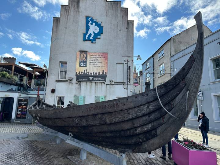 Full-scale wooden Viking longboat display set against pastel buildings under blue skies in Waterford.
