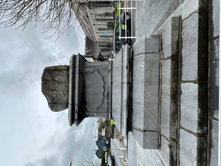 Weathered stone pedestal with an irregular rock mounted on top stands on a town pavement.