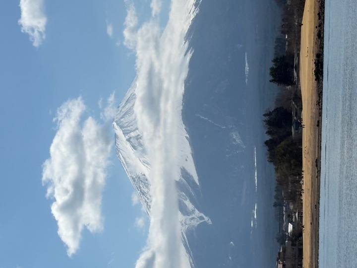 Snow-capped Mount Fuji rises above low clouds with a lake and rural foreground under a blue sky.