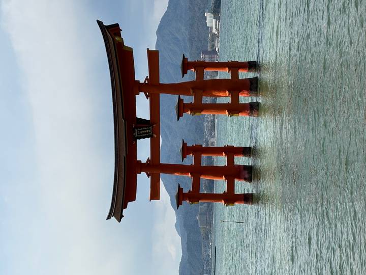 The iconic vermilion torii gate of Itsukushima Shrine standing in calm seawater with mountains behind.