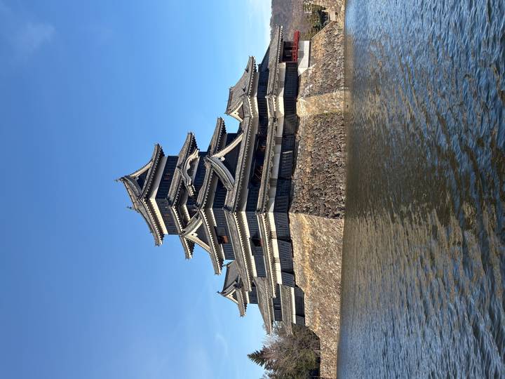 Dark-roofed Matsumoto Castle rises above its stone base and moat against a clear blue sky.