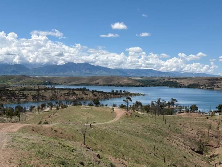 Large blue reservoir surrounded by rolling hills and distant mountain range under scattered clouds.