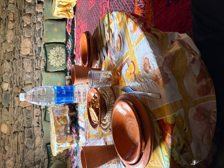 Simple meal setup with clay bowls, bread, tea glass and bottled water on a patterned plastic tablecloth.