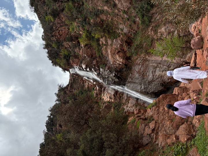 Two hikers approach a tall cascade flowing down rugged red cliffs amid green shrubs.