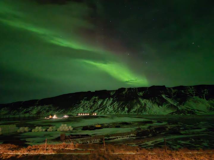 Brilliant green aurora borealis dances above snowy Icelandic mountains and farm lights