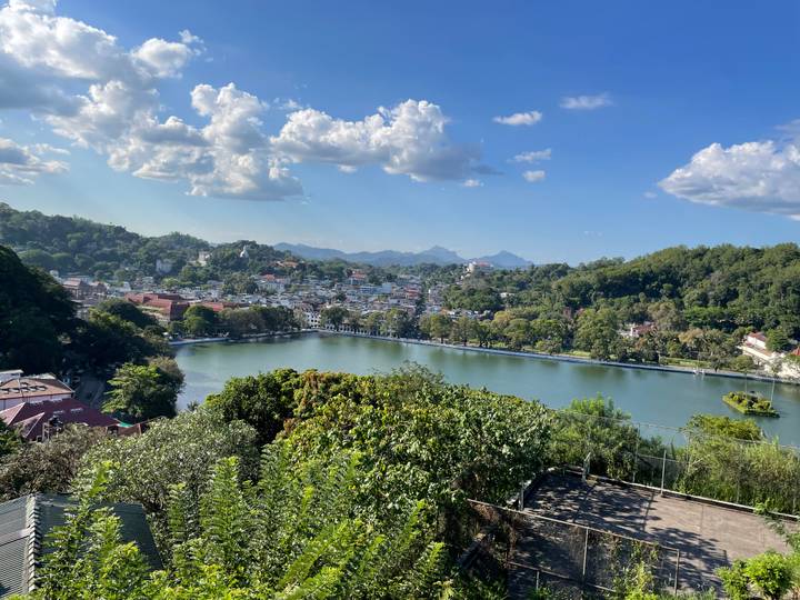 Scenic view over a tree-lined lake surrounded by a hillside city and distant mountains under a clear blue sky