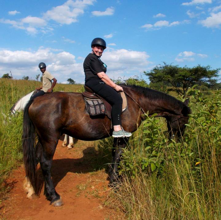 Two riders sit atop horses on a grassy trail under a blue sky in rural South Africa.