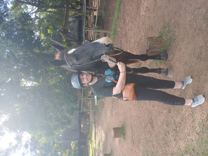 A woman wearing a riding helmet stands next to a dark horse in a shady paddock area.