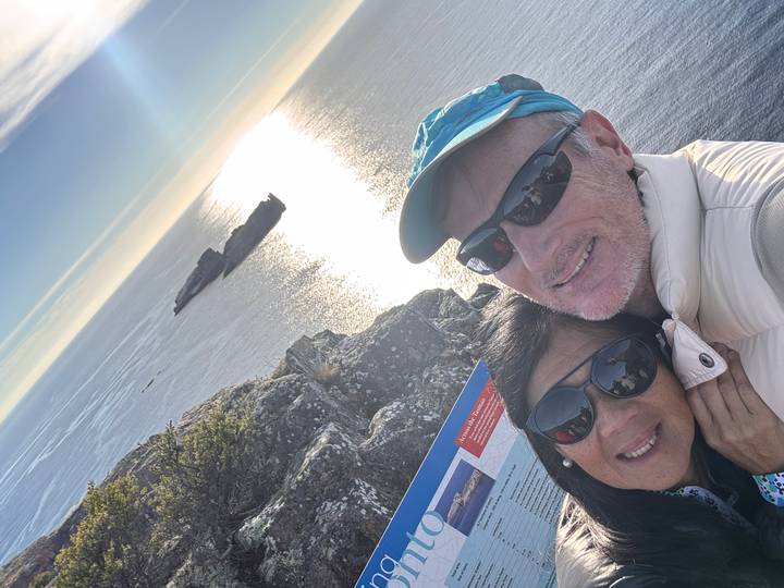 Couple takes a sunny selfie atop seaside cliffs with sparkling ocean and islands behind them.