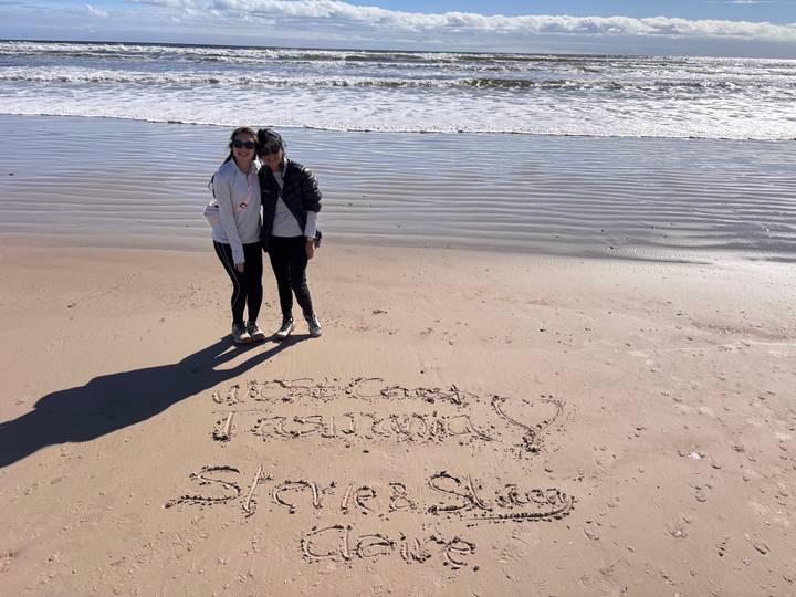 Two friends stand on a Tasmanian beach where words and a heart are written in the sand.