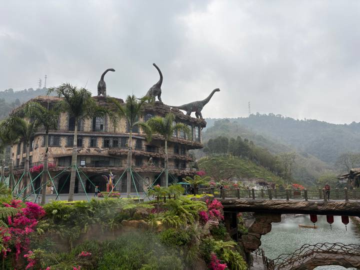 Themed building with dinosaur statues on roof, tropical plants, bridge and misty hills behind