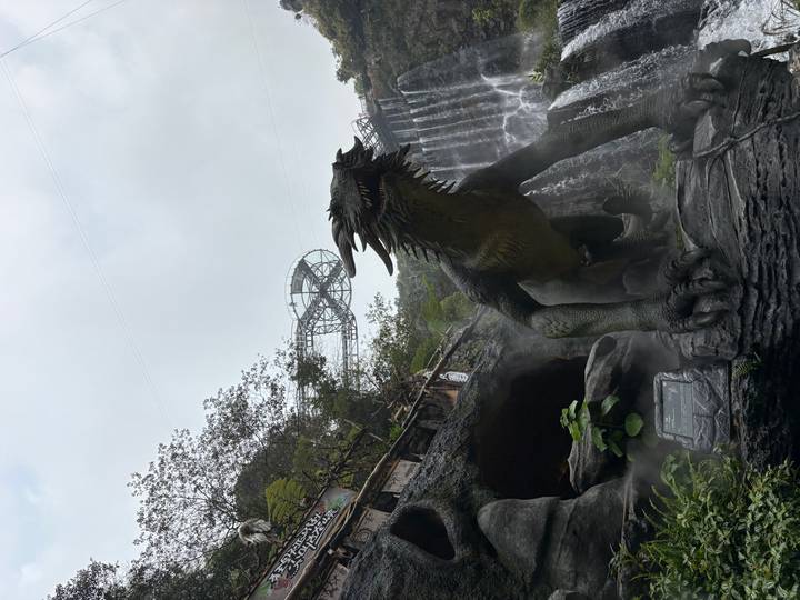 Large dinosaur statue in misty theme park with waterfall backdrop and observation deck