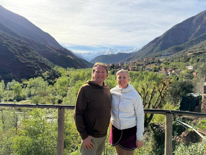 A smiling couple pose on a wooden railing with lush green valley and snowy Atlas peaks behind.
