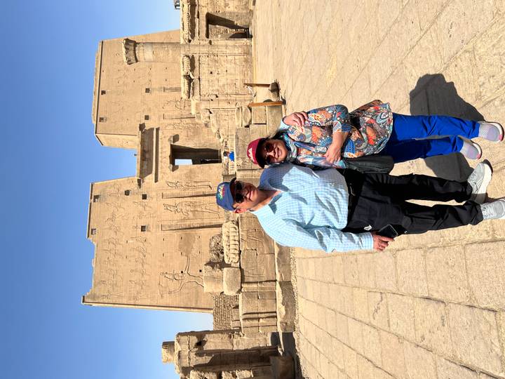 Couple stands before the sandstone pylons of Edfu Temple, hieroglyphs visible in the background.