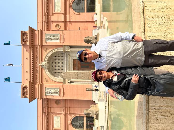 Visitors smile outside the pink-facade Egyptian Museum with ancient statues and a reflecting pool.