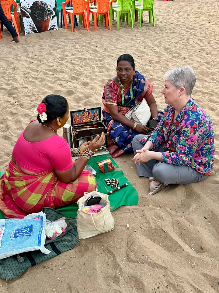 A local woman in a bright sari performs a cultural reading for a tourist seated on the sand with ritual items laid out.