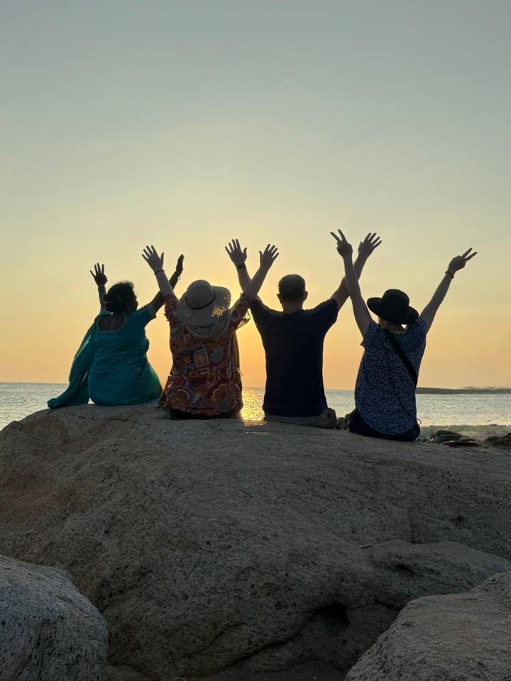 Silhouettes of four travellers seated on a seaside rock raise their hands toward a setting sun.