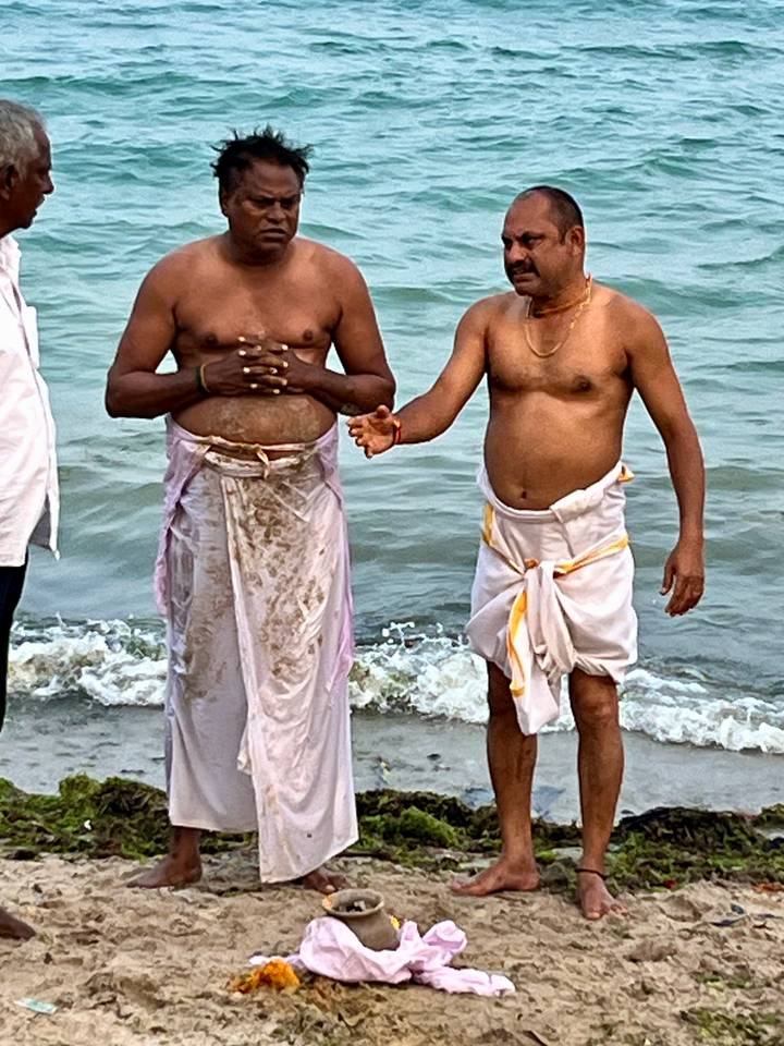 Two bare-chested men in traditional dhotis stand ankle-deep in the sea during a ritual dip.