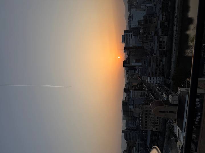 City skyline at dusk with the sun setting over low buildings and a jet contrail streaking the sky.