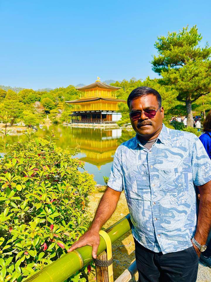 Man poses in front of the shimmering Golden Pavilion reflected in a tranquil pond.