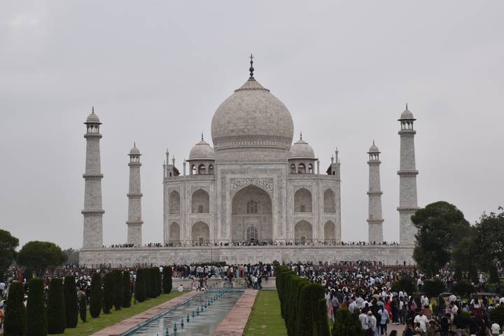 Front view of the Taj Mahal with large crowds gathered along the reflecting pool on an overcast day.
