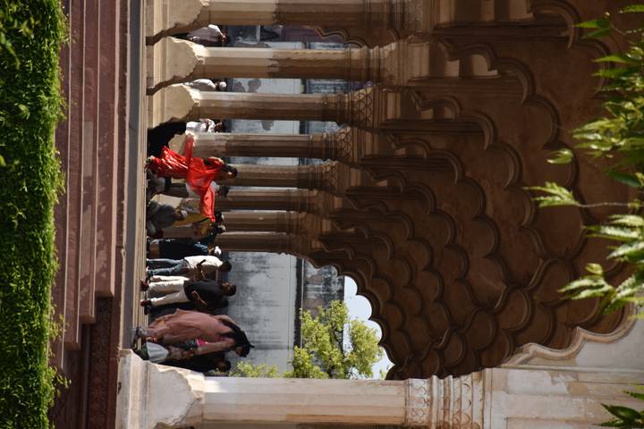 People move beneath a corridor of scalloped Mughal arches with a woman in a bright red dress twirling.