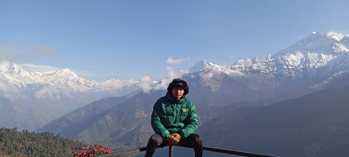 Young man in green down jacket sits on a railing with snow-capped Annapurna range behind.