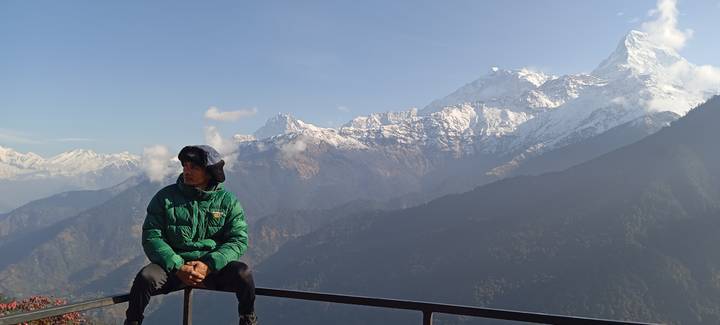 Traveller gazes to the side while seated on a balcony rail with panoramic snowy peaks.