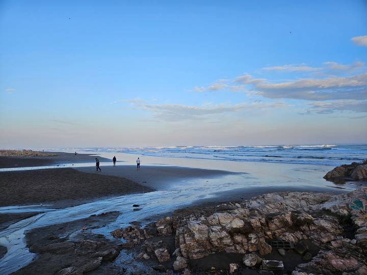Locals stroll along wide sandy beach at dusk with gentle surf.