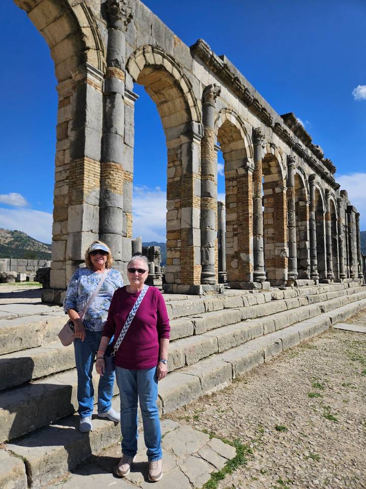 Two travelers pose among towering stone arches of ancient Volubilis ruins.