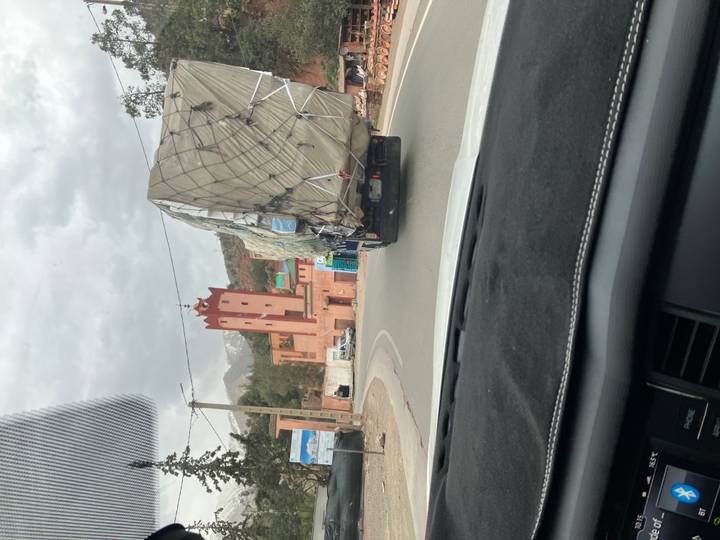 View through a car windshield of a winding mountain road with an overloaded truck and a red tower ahead under cloudy skies.