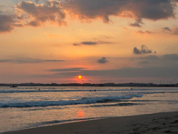 Orange-pink sunset over the ocean with small surfers riding gentle waves.