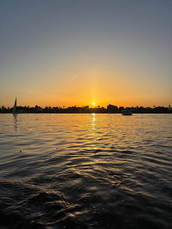 Golden sunset over a wide river with gentle ripples and silhouetted sailboats against a palm-lined distant shore.