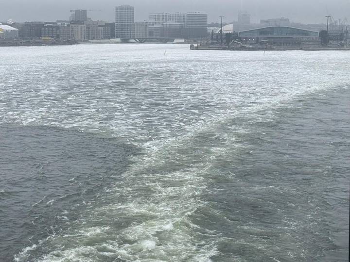 Choppy, partially frozen grey water with a foamy boat wake under an overcast sky and distant bridge.