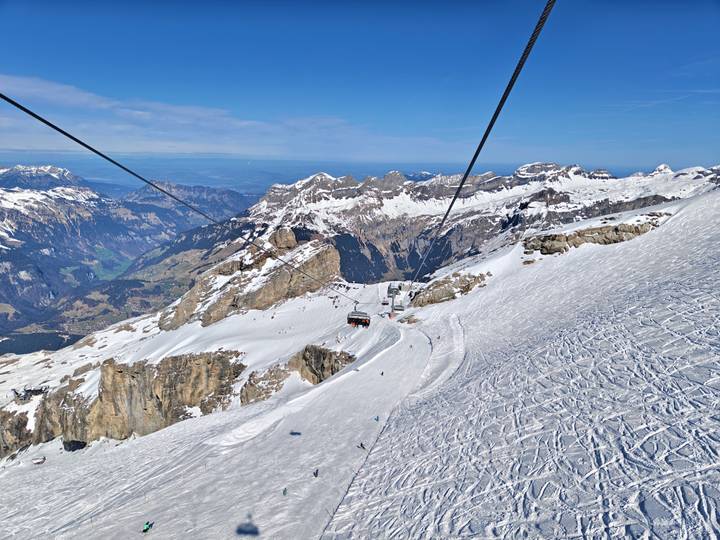 High-altitude view of snowy alpine peaks with a ski lift and cable cars descending toward a deep valley.