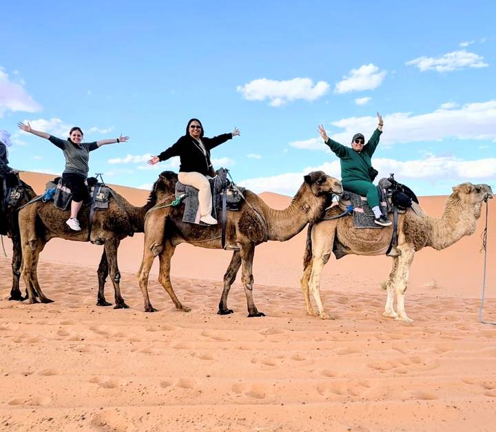 Three friends posing joyfully on camelback in the orange Sahara dunes under a blue sky