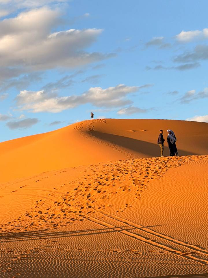 Golden sand dune with footprints leading to a lone figure on the crest while two people stand in conversation below