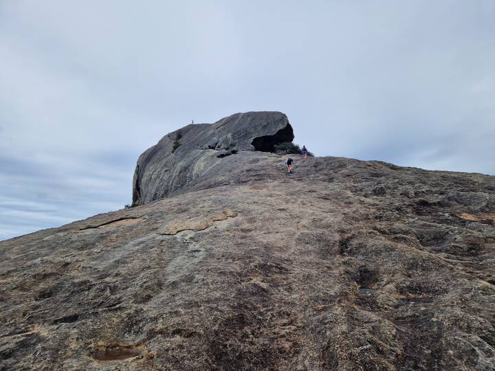 Two walkers ascending a massive granite outcrop under pale skies.
